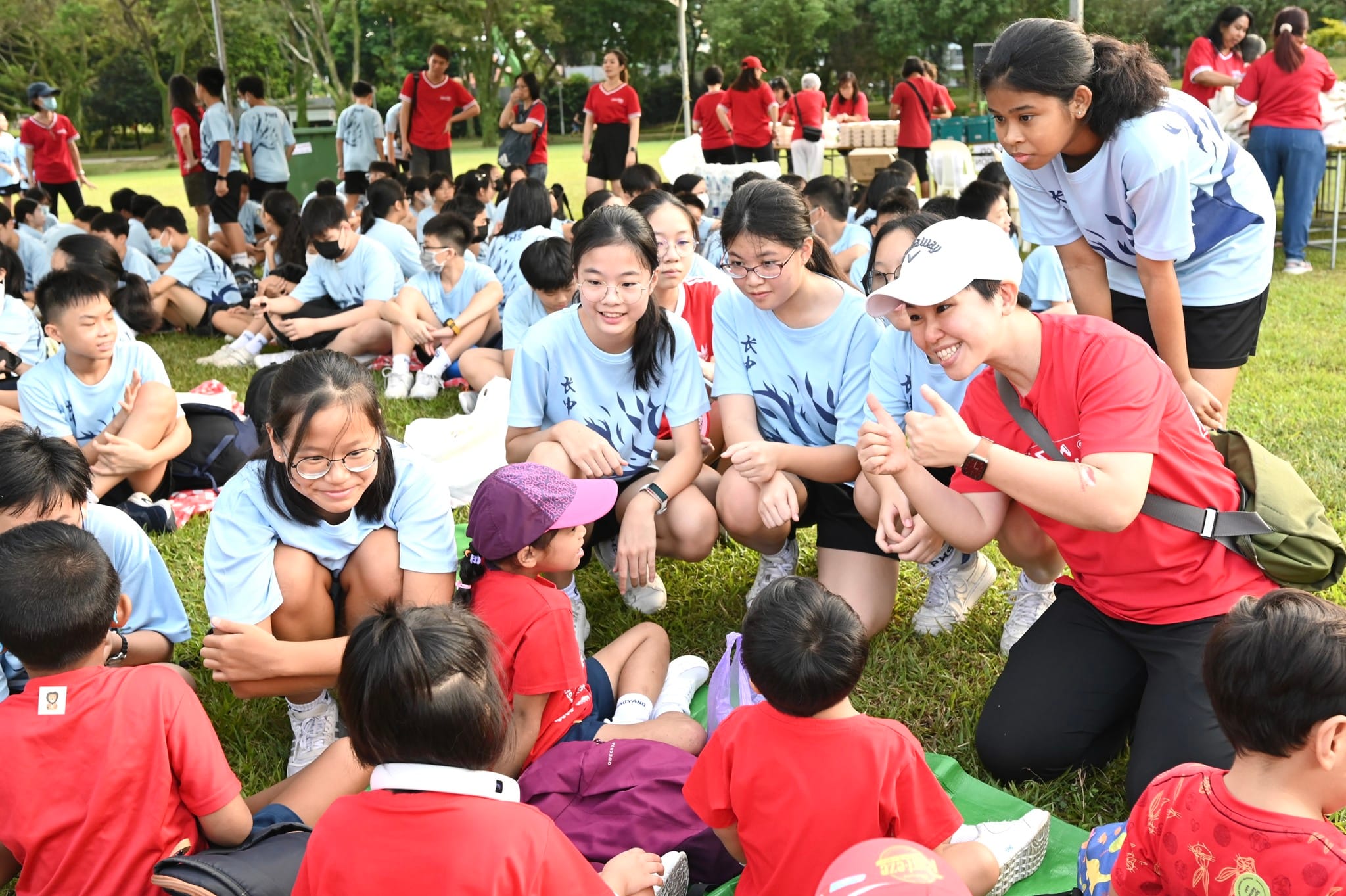 Large group of children and adults in blue and red shirts sitting on grass at an outdoor school event.